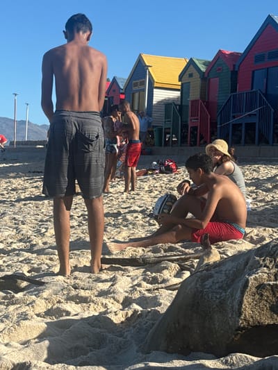 The image shows a beach scene in Muizenberg, South Africa, characterized by colorful beach huts in the background. Several people are present on the sandy beach. A shirtless man in striped shorts stands in the foreground with his back to the camera. In the middle ground, there are other individuals, including a man in red shorts. A woman in a sunhat is positioned near a shirtless boy. The environment consists of sand, beach huts, and a clear blue sky.