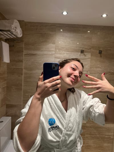 A young woman in a white robe takes a selfie in a bathroom with beige marble walls. She holds a blue smartphone and poses with one hand raised, displaying her red-spotted nails. The robe has a logo embroidered on the chest. A towel warmer with folded white towels is visible in the background, along with two downlights on the wall.