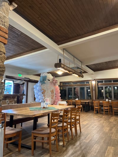 This image shows the interior of a restaurant, possibly in Smilyan, Bulgaria, given the context. The room has a high, vaulted ceiling with exposed wooden beams. A long wooden table is set for a celebration, with chairs on either side. A balloon arch in pastel pink and blue is positioned above the table, suggesting a baby shower or gender reveal event. The floor is made of light-colored wood planks. A window provides natural light, and a refrigerated display case is visible near the back of the room. The setting appears to be a private or semi-private dining area within the restaurant.