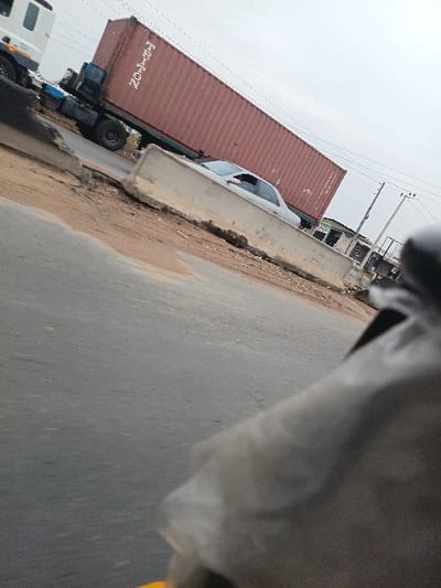 The image captures a roadside scene in Orifite, Nigeria, under an overcast sky. A white truck cab is visible in the upper left, attached to a long, red shipping container with the word "TRITON" inscribed vertically in white letters. To the right of the truck, a light-colored sedan is partially visible behind a damaged, light grey concrete barrier. This barrier spans horizontally across the mid-ground, separating a darker asphalt road in the foreground from the area containing the truck and car. A dirt and mud shoulder lies between the asphalt road and the concrete barrier. In the background to the right, utility poles with overhead wires and distant, indistinct structures are present. The perspective is from a low angle, with a blurred dark object and a small yellow element occupying the extreme lower right foreground. No individuals are visible.