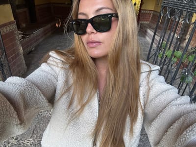 An upside-down selfie captures a woman with long blonde hair wearing black sunglasses and a cream-colored fuzzy jacket. She stands near a tiled area with a partially visible doorway and intricate metal railings with greenery. The background features a textured stone surface and parts of the architecture, giving an urban outdoor setting.
