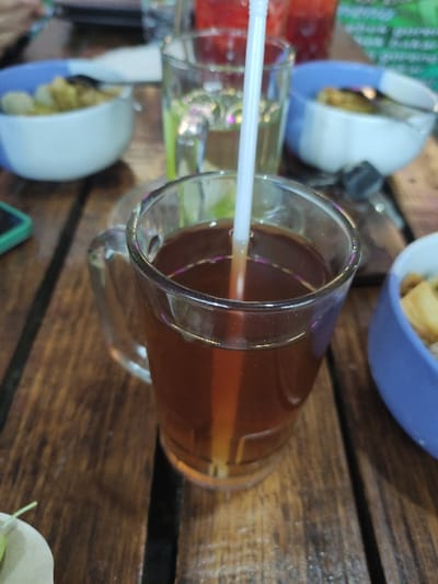 The image captures a close-up, overhead view of several drinks and snacks arranged on a rustic wooden table. The primary subject is a clear glass mug, filled with a dark amber-colored liquid, likely tea, into which a white plastic straw is inserted. The liquid shows a slight translucency.
To the left of the main mug, another clear glass with a lighter liquid, possibly water, is partially visible. Further left, a cup or glass containing a red-colored beverage can be seen. Scattered across the table are several bowls. One prominent blue and white bowl holds a serving of small, light-colored, fried food items, resembling crackers or chips. A similar bowl is partially visible in the upper background.
The setting appears to be a casual dining or relaxation area, likely outdoors or in an establishment with a natural aesthetic, given the sturdy wooden table. The table surface is composed of distinct wooden planks with visible gaps. The overall arrangement suggests a social gathering or a moment of refreshment.
The lighting in the image is soft and even, suggesting either an overcast daytime environment or an indoor setting with diffused light. No specific time of day is discernible, but it does not appear to be nighttime. The focus is sharp on the central drink, with the background elements slightly blurred. There is no visible text identifiable on any of the objects or in the background.