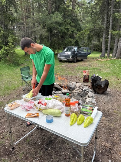 In an outdoor setting, likely a forest or wooded area near the city of Kosovo, Bulgaria, a young man is preparing food at a campsite. He is wearing a green t-shirt and black shorts, and is actively chopping a zucchini on a white plastic cutting board. In front of him is a foldable picnic table laden with various food items, including packaged meats, fruits, vegetables, a can, and bottles of sauce and oil. Behind the table, a campfire crackles, surrounded by rocks. A dark-colored car with its doors open is parked in the background, partially obscured by trees. A green folding chair sits to the left of the man, and a bag of charcoal is placed near the campfire. The environment is characterized by a forest floor covered in dirt and pine needles, with tall trees and green undergrowth visible.