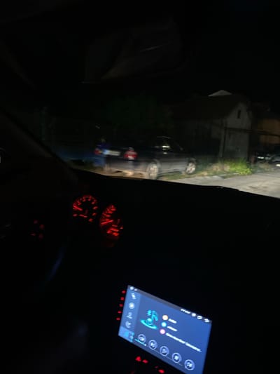 The image depicts the interior of a car at night, with a view through the windshield. The dashboard features illuminated red gauges, indicating speed and RPM. A digital display screen, likely a car infotainment system, is visible in the foreground, showing a music interface with icons and text. Outside the car, a dark street scene is visible. To the left, a black car is parked or moving slowly, with its headlights illuminating the road and adjacent greenery. Further in the background, buildings are dimly lit, suggesting a residential area. A person in a blue shirt is standing near the parked car. The scene is consistent with a street in the city of Yasen, Bulgaria.