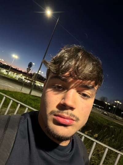 The image presents a close-up, selfie-style portrait of a young male, estimated to be in his late teens or early twenties, captured outdoors at dusk or night in Penafiel, Portugal. The subject possesses dark, wavy hair, distinct eyebrows, a mustache, and a short beard. He wears a dark, possibly navy, crew-neck t-shirt, with the strap of a dark backpack visible over his left shoulder. His gaze is directed forward, into the camera lens.
The background reveals a transitional environment. The sky is dark blue, indicating evening or night, with a faint crescent moon visible in the upper right quadrant. Artificial illumination is a dominant feature, with a bright street lamp positioned directly behind the subject, generating a starburst effect. Multiple additional streetlights are visible, illuminating a paved area, which appears to be a road or parking lot, where several vehicles are faintly discernible. Buildings with illuminated interiors/exteriors are present in the mid-distance, suggesting a commercial or developed urban area. Green grass and scattered vegetation are visible along the paved surface. A metal railing, consisting of horizontal bars, occupies the lower foreground, partially obscured by the subject. Traffic signs, including a circular sign with a red border, are present in the mid-ground.