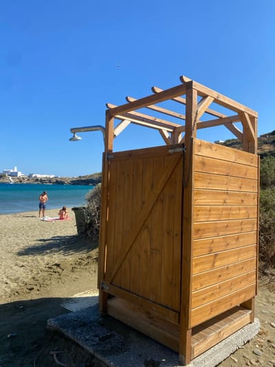The image displays a sunlit beach scene in Φάρος, Greece. In the right foreground, a freestanding outdoor shower enclosure is positioned on a concrete slab base on light-brown sand. The structure is constructed of light-brown wooden planks, featuring a hinged door with a diagonal brace on its left side and an open wooden framework at the top. A silver-toned metal shower head on an L-shaped arm extends from the left side of the enclosure. A slatted wooden platform is visible at the shower's base.
In the midground, on the sandy beach near the water's edge, two individuals are present. A male, wearing blue swim trunks, stands facing left, while a female lies on a pink towel to his right. A dark container is partially visible near the female, partially obscured by sparse vegetation. The background features a body of clear turquoise water, transitioning to deeper blue. Beyond the water, a rocky hillside rises, dotted with white Cycladic-style buildings, including a distinct white building with a domed roof, likely a church or chapel, on the distant shoreline. A small, dark object is discernible in the upper portion of the clear blue sky. The scene is illuminated by bright sunlight, casting defined shadows.