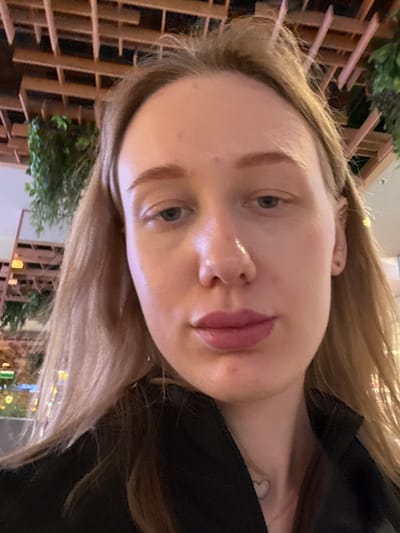 A close-up, rotated selfie of a fair-skinned woman with light brown hair. She is wearing a black top. The background appears to be an outdoor setting in Ol'ginskaya, Russia, suggested by the presence of a lattice-style fence with greenery and distant lights.