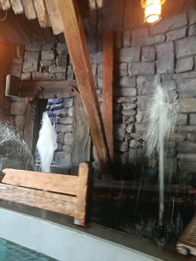 The image depicts an indoor scene with a rustic, stone-like wall and wooden elements, reminiscent of a mine or old structure. Water is actively spraying from multiple points, creating dynamic water features. One prominent water jet erupts on the right side of the frame, while another, more dispersed spray, is visible on the left. In the center, a tall, white plume of water shoots upwards through an opening in the stone wall. A wooden bench or trough is situated in the foreground, appearing wet from the water. Wooden beams are visible overhead and supporting the structure. A light fixture is present in the upper right corner, emitting a warm glow. The overall impression is of a themed attraction, possibly within a park or entertainment venue. The location in Brühl, Germany, suggests it could be within the Phantasialand theme park, which features attractions with similar thematic elements.
