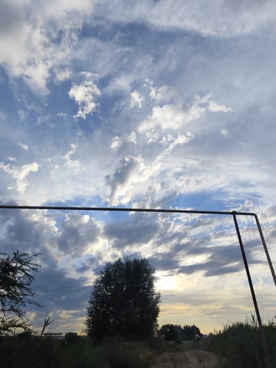 This is a landscape photograph taken at sunset in Dolan, Kazakhstan. The image features a dramatic sky filled with a mix of light and dark clouds, with patches of blue sky visible. The sun is setting, casting a warm glow on the clouds and creating a vibrant atmosphere. In the foreground, a rudimentary metal structure, possibly a part of a playground or sports equipment, is visible. Silhouetted against the sky are trees and tall grass, suggesting a rural or semi-rural setting. A dirt path or road winds through the scene, leading the eye towards the horizon. The overall mood is peaceful and serene, characteristic of a rural sunset. There is no visible text in the image.