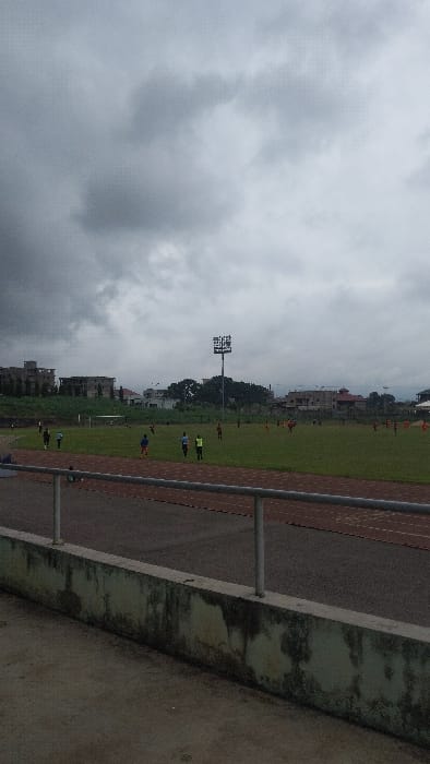 The image shows a view of a soccer field in Buea, Cameroon, taken from a nearby elevated platform. The coordinates provided place the scene at the Buea Stadium, which is located on the outskirts of the city. The field is green and surrounded by a low wall. There are a few people on the field, some standing, some walking, and some appear to be playing soccer. There is a single, tall, gray metal light post in the center of the field, with multiple lights on top. The sky is overcast with gray clouds. The scene is captured from behind a low wall, with a metallic railing in the foreground. The ground is concrete, partially covered with moss. The view is taken from the top of the concrete bleachers on the side of the field.