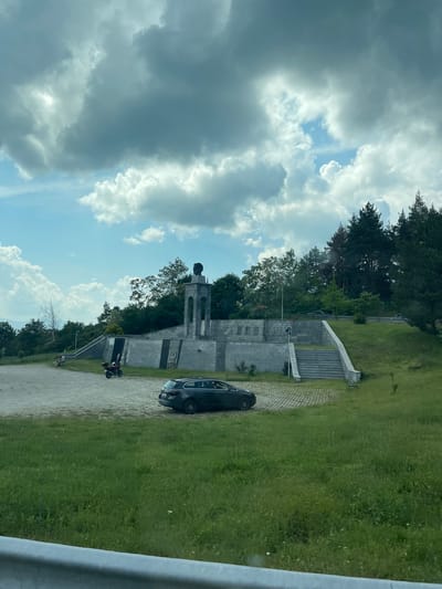 The image depicts an outdoor scene in Bunovo, Bulgaria, showcasing a concrete monument complex situated on a hillside under a partly cloudy sky. In the immediate foreground, a silver-colored guardrail is visible, positioned above a strip of unkempt green grass containing various weeds and small white flowers.

The midground features a large, irregularly paved area of light-colored interlocking pavers. A dark grey station wagon, identifiable as a wagon-style vehicle, is parked on this paved surface towards the right, presenting its rear profile. To the left of the car, a red motorcycle is parked. One individual is visible seated on a bench in the far left section of the paved area, facing towards the distant landscape.

The central subject is a multi-tiered monument constructed from light grey concrete or stone. The lower level consists of a retaining wall with several carved rectangular panels and integrated staircases leading upwards. The upper level features a more elaborate retaining wall with additional decorative elements, possibly relief carvings or plaques. At the apex of the monument complex stands a tall, columnar structure with open arched sections beneath a prominent bust of a male figure, facing towards the viewer's left.

The background is dominated by dense vegetation. A mix of deciduous and coniferous trees covers the hills surrounding the monument. A thick forest of pine trees is particularly prominent on the right side of the image, extending upwards. A paved road with a guardrail is visible winding through the trees on the hillside behind the monument, with a dark-colored car present on it. Distant, hazy mountains are faintly discernible on the far left horizon beneath a clearer portion of the sky. The sky above is characterized by large, dark grey cumulus clouds intermixed with patches of bright blue sky and smaller, whiter clouds.