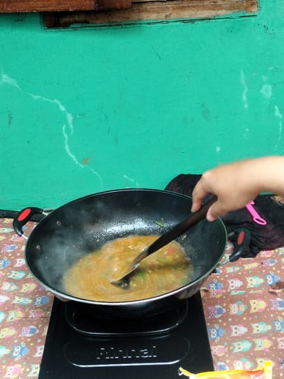 The image shows a close-up view of a person cooking in what appears to be a home or sheltered outdoor setting in Leuwiliang, Indonesia. A right hand, with a wooden spatula, is actively stirring a simmering, orange-brown liquid or sauce within a black wok. Visible steam rises from the wok, indicating heat and active cooking. The wok rests on a black Rinnai-branded electric stove.
The cooking surface underneath the stove is covered with a patterned fabric featuring repeating cartoon owl designs in various colors. The background is dominated by a brightly colored teal or turquoise wall, which displays several white, hair-thin cracks and a faint, darker discoloration. A rustic wooden plank or beam runs horizontally across the top of the wall, adding to the informal aesthetic. To the right of the wok, a crumpled piece of dark fabric is visible, alongside a bright pink utensil, possibly a pair of tongs. A small section of a yellow package is also seen at the bottom right corner of the frame. The scene suggests a daytime activity.
