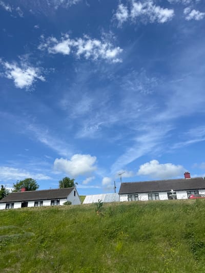 The image depicts a rural Irish scene in Donegal, with a focus on a bright blue sky filled with wispy clouds and some puffier white clouds. To the right, a white-walled house with a dark tiled roof and a red chimney runs along the edge of the frame. A tall, overgrown grassy field with scattered yellow wildflowers borders the house. Another section of a white building, possibly an extension or a separate structure, is visible at the bottom. The image appears to have been taken during the daytime, under fair weather conditions. There is no visible text.