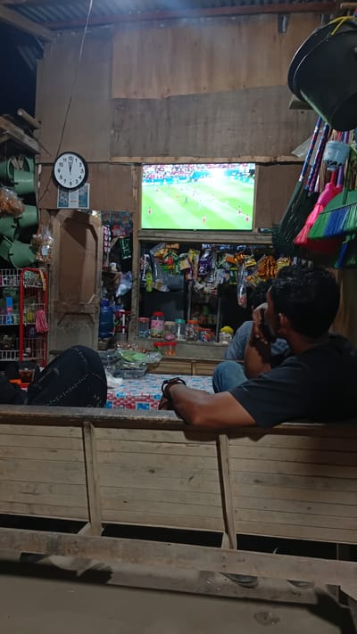 This media file depicts a dimly lit, rustic indoor scene, likely a small shop or stall in Matangcut, Indonesia. The primary subjects are two individuals and various objects related to a shop's inventory.
One person, seen from the side and from behind, is wearing a dark t-shirt and jeans and appears to be looking down at something in their hands, possibly engaged in a task or examining an item. The other person is mostly obscured, wearing a dark headscarf and clothing, and is also looking downwards.
Prominent objects include a television screen displaying a soccer match with a bright green field and players. Shelves are stocked with a variety of packaged snacks and drinks, typical of a small convenience store. Other items include buckets, brooms, a clock showing approximately 9:10, and what appear to be cleaning supplies. The setting is constructed from rough wooden planks, giving it a worn and makeshift appearance. The overall lighting is dim, suggesting it is either evening or the interior of a poorly lit structure. There is no visible text in the image. The atmosphere appears to be casual and everyday, with the focus on the television and the shop's wares.