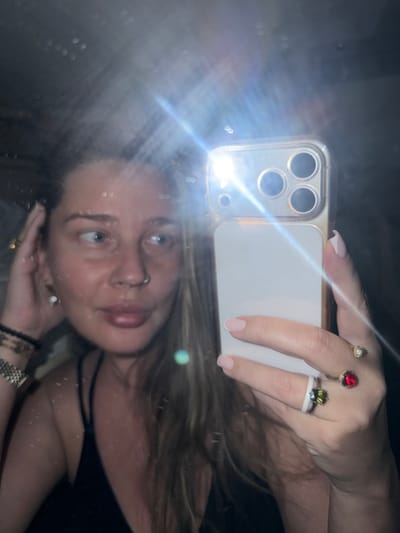 A selfie in a mirror shows a fair-skinned person with blonde hair, blue eyes, and a nose ring. They are holding a white smartphone with three visible camera lenses. The phone's flashlight is on, causing a bright glare. The person is wearing several rings on their right hand: one with a red stone, one with a yellow stone, and one with a green stone atop a white band. They are wearing bracelets on their left wrist. The reflection shows the person wearing a black top. The background is dark and indistinct. The location is not identifiable as Krasnyi Yar, Russia from the provided image.
