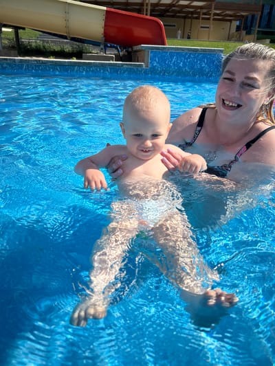 In the city of Yubileiny, Russia, a woman and a toddler are in a swimming pool. The woman, with blonde hair, is wearing a black swimsuit with a colorful pattern. She is holding the toddler, who has light hair and blue eyes, in the water. The toddler is wearing a diaper and appears to be enjoying the swim. The water in the pool is bright blue and appears clear. In the background, there is a red and yellow water slide and some shaded seating areas. The scene suggests a recreational setting, possibly a water park or a public pool in Yubileiny.