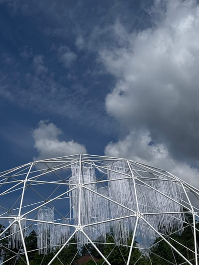 This is a low-angle, wide shot of a geodesic dome sculpture against a blue sky with scattered clouds. The sculpture, made of white metal framing, appears to be covered in a transparent or white material that hangs in vertical strands, creating a textured surface. The dome is partially obscured by dark green foliage on the right side of the frame, suggesting it is located outdoors, possibly in a park or garden. The lighting indicates it is daytime, with the sun casting some shadows on the clouds, suggesting it might be late morning or early afternoon. The overall mood is serene and contemplative, with the vastness of the sky contrasted by the intricate structure of the sculpture. No people are visible, and there is no visible text. The location context provided is Yunost', Russia.
