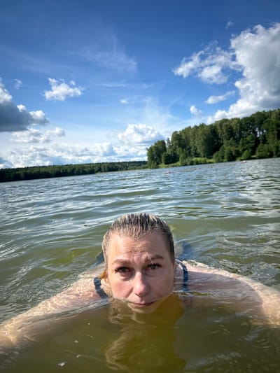 A woman is swimming in a lake on a sunny day with clouds in the sky. The lake is surrounded by a dense forest. The water is slightly choppy, and sunlight reflects off the surface. The woman is looking towards the camera, with her head and shoulders visible above the water. She is wearing a dark swimsuit. The image appears to be taken from a low angle, close to the water's surface. There are buoys visible in the distance, marking a swimming area. The location is the city of Yubileyny, Russia.
