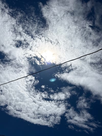 A low-angle, full shot captures a dark blue sky dotted with scattered white cumulus clouds. The sun is partially obscured by clouds in the upper center of the frame, creating a bright halo effect and a lens flare in the lower center. A thin, black power line stretches diagonally from the bottom left to the upper right across the sky. The scene is devoid of any people or discernible landmarks, suggesting an outdoor setting, possibly rural or suburban. The image lacks any context indicating a specific location within the Town of Cheektowaga, United States of America.