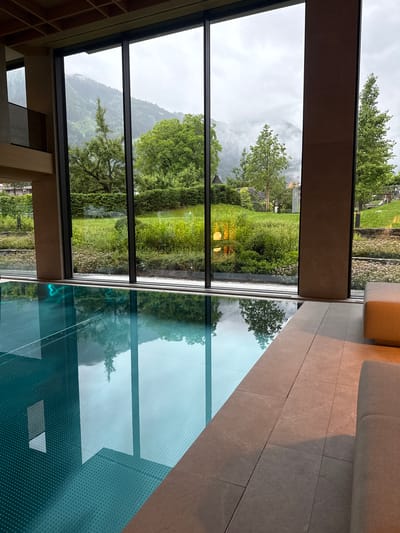 The image depicts an indoor swimming pool area within a modern resort in Mayrhofen, Austria. Large, floor-to-ceiling windows offer a panoramic view of lush green gardens, trees, and mist-covered mountains in the background. The pool itself is a striking turquoise color, with a textured surface visible beneath the clear water. Reflections of the surrounding trees and the cloudy sky are visible on the water's surface. To the right of the pool, the floor is tiled with large, light-colored tiles, and a beige upholstered sofa is partially visible. The interior architecture features a wooden ceiling with a grid pattern and stone pillars. The overall scene conveys a sense of tranquility and luxury, with the natural landscape serving as a prominent backdrop.