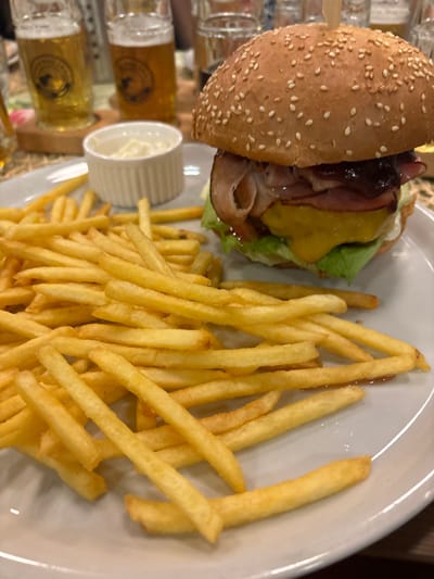 A sesame seed bun hamburger with bacon, cheese, and lettuce on a white plate. Accompanying the burger are fries and a small ramekin of white sauce. Several small glasses of what appears to be beer are on the left side of the plate. The plate is tilted in the shot, with the beer glasses on the lower-left side of the photo.