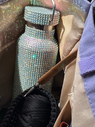 The image displays a close-up of a bedazzled water bottle and crochet supplies inside a bag. The water bottle is covered in numerous small, reflective rhinestones that create a shimmering effect, enhanced by light glinting off their surfaces and projecting colorful reflections onto the surrounding fabric. A crochet hook with a wooden handle, etched with the word "Gamma," is positioned in the foreground, with its metal hook engaged in black yarn. The yarn is wound into a thick, coiled shape. The bag appears to be made of different fabrics, including a beige material and a light purple fabric with a textured band. The scene is lit in a way that highlights the sparkle of the bottle and the textures of the materials. There is no discernible information in the image to indicate a specific location in Сухарэўскі сельскі Савет, Belarus.
