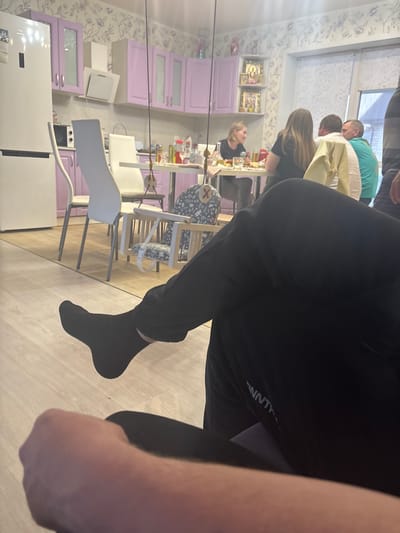 The image shows a group of people in a residential kitchen, likely located in Nestiukovo, Russia. The kitchen has lavender cabinets, a white refrigerator, and white chairs around a dining table. Several people are gathered at the table, appearing to be engaged in a meal or conversation. The foreground is dominated by a person wearing black pants and a black sock on their foot. The focus is somewhat distorted, with certain elements appearing closer than others, suggesting a close-range perspective.