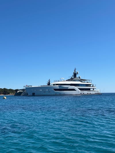 A large, modern white superyacht is anchored in the clear blue waters off the coast of La Maddalena, Italy. The yacht features multiple decks, sleek lines, and dark-tinted windows. A smaller, darker hull yacht is docked alongside the superyacht, partially obscured. In the background, a tree-covered island with a small beach is visible. The sky is a clear, bright blue. A yellow buoy floats in the foreground, just to the left of the superyacht. The water is calm with gentle ripples.