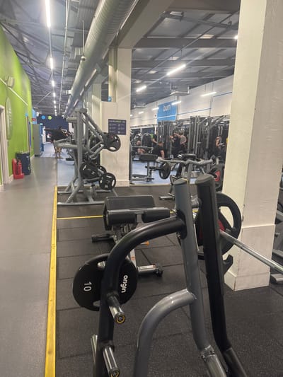 The image shows the interior of a gym in Sheffield, United Kingdom. The space is dominated by weightlifting equipment, including a lever row machine with "Origin" branded weight plates (10kg) in the foreground. Other weight machines are arranged in rows, with individuals using them further into the room. Overhead, there is a large ventilation duct and fluorescent lighting. The floor is a mix of gray mat and green and blue cushioned areas. There is a yellow safety line along the edge of the gray mat. Some personal bags sit on the ground along the green and blue cushioned areas.