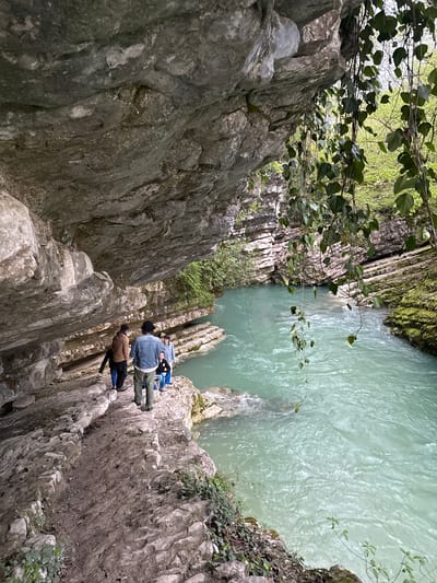 The image depicts a river gorge in Lebache, Georgia. The gorge walls are composed of jagged, layered rock formations, dropping sharply to a turquoise-colored river below. Two individuals are positioned on the edge of the gorge. One is wearing a denim jacket, dark pants, and blue shoes and is lying with their head towards the water. A second person is standing beside them in a brown jacket, black pants, and blue shoes. The viewpoint is looking down into the gorge, capturing both the rock structure and the water. Some foliage and trees are visible along the top edge of the gorge.