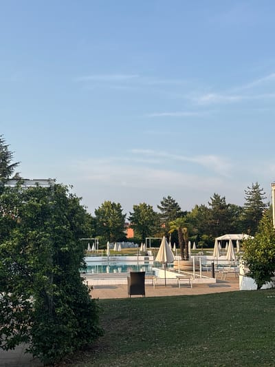 This is an outdoor daytime photograph taken in Pieve Emanuele, Italy, likely at a resort or recreational facility. The scene is dominated by a swimming pool area with several lounge chairs and white umbrellas. The pool itself is partially visible and appears to be filled with clear water. Surrounding the pool area are lush green trees and grass. In the background, there are buildings and more trees, suggesting a suburban or semi-rural setting. The sky is a clear, pale blue with a few wispy clouds. There are no people visible in the image. The lighting suggests it is either early morning or late afternoon, with a soft, diffused light.
