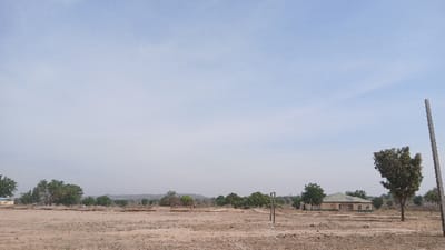 The image displays an arid, open landscape in Bashar, Nigeria, under a clear, hazy blue sky. The foreground and midground consist of dry, light-brown earth with minimal ground cover. Scattered green-leafed trees are distributed across the midground and background. A tall, grey vertical pole occupies the right foreground. Towards the center-right midground, two shorter, dark wooden posts are visible. In the background, a cluster of light-colored buildings with dark or green roofs is present, including one distinct light yellow/tan structure with a green roof. No individuals or discernible activities are observed.
