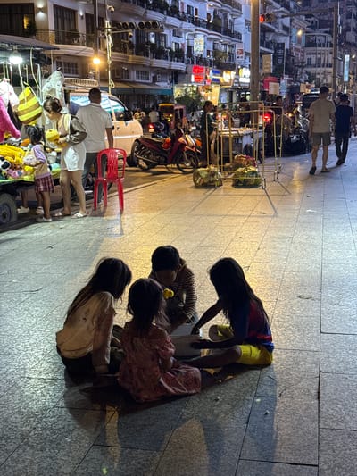 The image depicts a street scene at night in Khan Daun Penh, Cambodia, likely a commercial or pedestrian area given the presence of shops and street vendors. In the foreground, four young children are seated on the pavement in a circle, appearing to be interacting with a white tub. The children are dressed in casual, varied clothing. To the left, a street stall displays a variety of stuffed toys and bags. A woman and a man are visible near the stall, with the woman holding a yellow stuffed toy and the man standing behind her. A red plastic chair is placed next to them. Further back, the street is lined with multi-story buildings with illuminated windows and signage, suggesting shops or residences. Several motorcycles are parked or in motion, and other pedestrians are walking on the sidewalk. The lighting appears to be a mix of streetlights, shop lights, and building illumination, casting shadows on the paved ground. The overall atmosphere suggests a bustling urban environment.