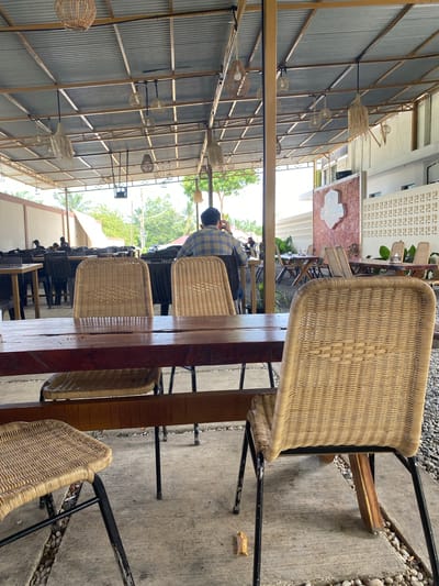 The image shows an outdoor seating area of a restaurant in Aluepiet, Indonesia. The area is covered by a corrugated metal roof supported by a metal frame. Several woven pendant lights hang from the ceiling. In the foreground, a wooden table is visible with woven chairs around it. In the background, a person wearing a plaid shirt sits at a table, facing away from the camera. Other tables and chairs are arranged in the seating area, and trees and foliage are visible in the distance. The ground is a mix of concrete and gravel.