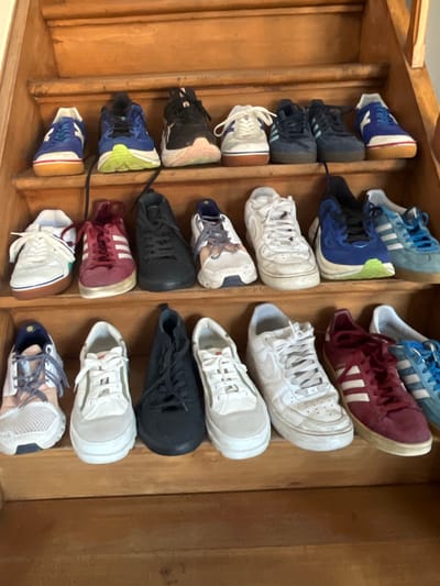 The scene, located in Foetz, Luxembourg, depicts an indoor environment featuring a light to medium brown wooden staircase. Multiple pairs of athletic footwear are systematically arranged on the steps.
On the topmost visible step, three pairs of shoes are present: a blue and white low-profile sneaker positioned on the left, a dark blue and black running shoe with a neon green sole in the center, and a black running shoe with a pink and white sole on the right.
The second step from the top holds five pairs of shoes. From left to right, these include: a white and red classic athletic shoe with a brown sole, a dark red sneaker with white stripes, a black casual sneaker, a white casual sneaker resembling a Nike Air Force 1 style, and a light blue sneaker with white stripes.
The third step from the top also displays five pairs of shoes. From left to right: a light-colored mesh running shoe with subtle blue accents, a white casual sneaker resembling a Nike Air Force 1 style, a white casual sneaker, a black casual sneaker, and a dark red sneaker with white stripes.
The fourth visible step contains three pairs of shoes. From left to right: a white casual sneaker, a blue and white athletic shoe, and a light blue sneaker with white stripes.
In total, sixteen pairs of athletic shoes, varying in color and style from running shoes to casual sneakers, are displayed across four distinct steps of the wooden staircase. No individuals or active interactions are observed within the frame.