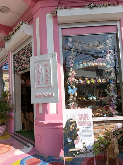 The image shows a brightly colored storefront in Puerto Plata, Dominican Republic. The building is painted pink with white vertical stripes. Above the entrance, artificial flowers are draped along a white awning. A sign that reads "HAND MADE AVARCA" is mounted on the wall. The storefront window displays several pairs of shoes arranged on shelves, decorated with artificial flowers and butterflies. A poster in front of the window features a woman crafting espadrilles, with text that reads "LIVE SHOE-MAKING in progress." To the right of the poster, a straw donkey statue is adorned with flowers. The ground in front of the store is painted with colorful geometric patterns.