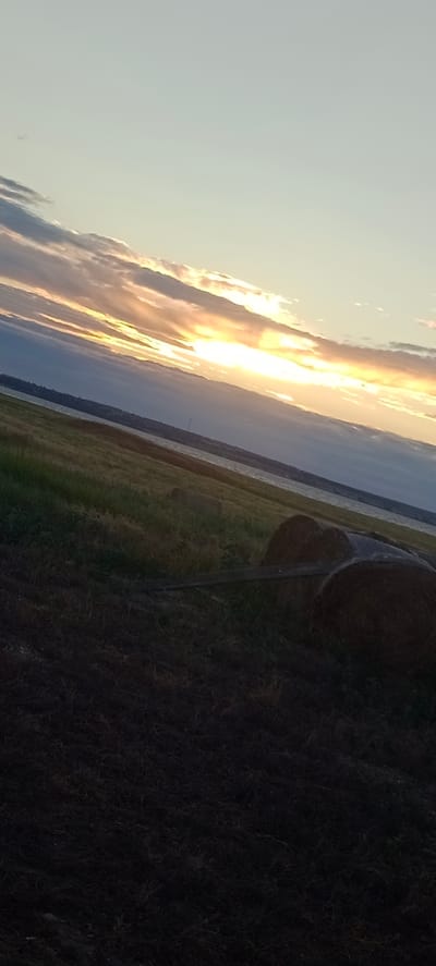 This is a photograph taken at sunset in Peskovatka, Russia. The image is horizontally aligned and shows a landscape with a body of water in the middle ground and a field in the foreground. The sky is filled with clouds, and the sun is setting, casting a warm glow across the scene. The water is calm, and there are a few boats visible on the water. The field is covered in grass and hay bales. There are no people or animals visible in the image. The overall mood of the image is peaceful and serene.