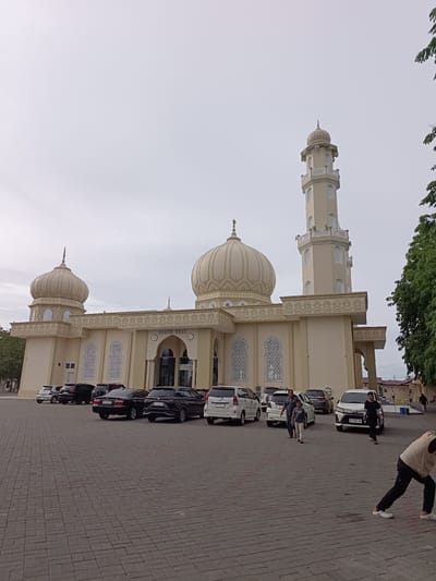 The image depicts the Baitul Huda Mosque in the city of Mee Tanjong Usi, Indonesia, under a cloudy sky. The mosque features a prominent minaret and several domes, with light-colored walls and decorative latticework on the windows. A paved courtyard in front of the mosque is occupied by several parked cars, including sedans and SUVs. People are visible in the courtyard, some walking and others standing near the cars. The overall scene suggests a public gathering or an active day at the mosque.