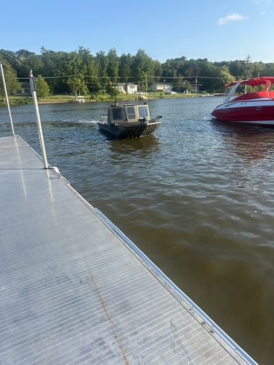 An outdoor, eye-level shot captures a lake with two boats and a dock. On the left, a metal dock juts into the frame. In the center of the lake, a small, dark green boat with a tan cabin and a person inside is moving away from the dock. To the right, a red and white boat with a red canopy is docked. The background features a tree line with houses and a clear blue sky. The scene is set in Algonquin, United States of America.