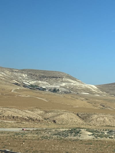 The image shows a landscape in al-Qasoum Regional Council, Israel. The scene features a clear blue sky occupying the left portion of the frame. The right side depicts a mountainous terrain with varying textures and colors, ranging from light beige to dark gray. A village or settlement is nestled within the mountain, characterized by a collection of buildings and green vegetation. A road runs alongside the base of the mountain, and a single vehicle, appearing red, is visible on the road. The landscape is arid and sparsely vegetated.