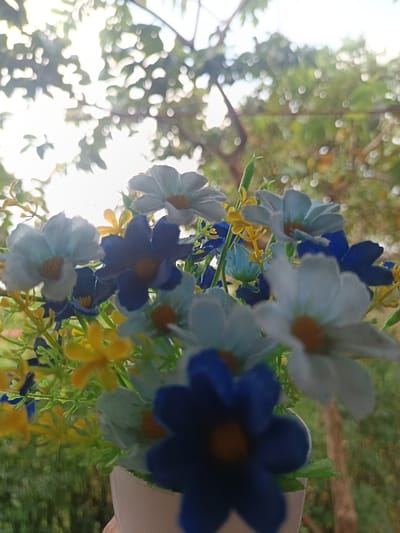 A close-up photograph, taken from a low angle, depicts an arrangement of artificial flowers held in a white container, set outdoors in Ouderkerk aan de Amstel, Netherlands. The bouquet features numerous small fabric flowers in white, light blue, dark blue, and yellow, each with an orange or yellow center. The white container is partially visible at the bottom center of the frame. The background is heavily blurred, displaying green foliage and tree branches, with bright natural sunlight filtering through, creating prominent bokeh effects. No identifiable individuals are present.