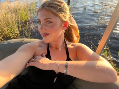 A woman is photographed at eye level, reclining with crossed arms, wearing bracelets and rings. She is wearing a black top and a gold choker necklace with a gem. Her hair is styled, and she has on hoop earrings. The background shows water, vegetation, and part of what appears to be a boat frame, suggesting a location near a body of water. The scene is brightly lit with sunlight. No specific location information within Victoria Falls, Zimbabwe, can be determined from the image.