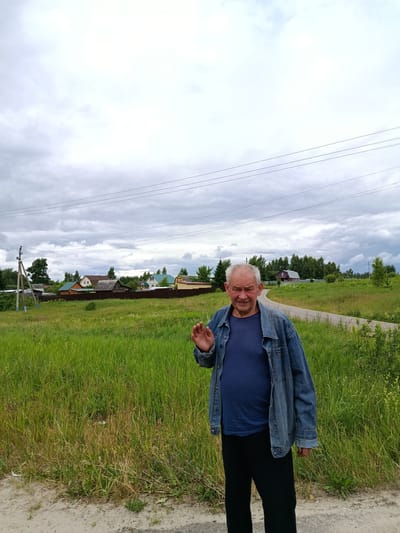 In the city of Gribchikha, Russia, a man with gray hair and a smiling face waves to the camera while standing in front of a rural landscape. He is wearing a blue t-shirt and a denim jacket, with black pants. The background features a dirt road curving through a field of tall green grass and a cluster of houses with various roof colors, suggesting a residential area. Trees and power lines are visible in the distance, under a cloudy sky.