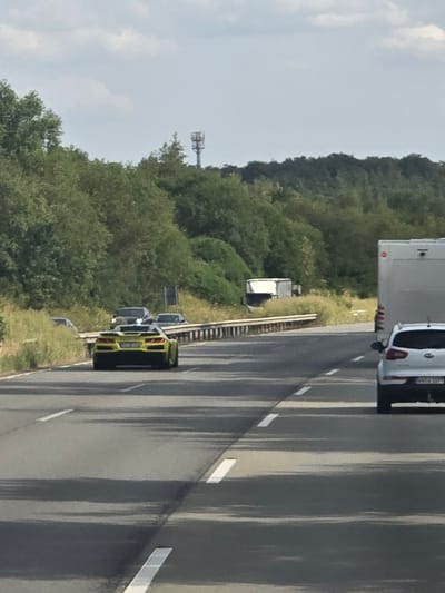 The image depicts a highway scene, likely in or near Siershahn, Germany, given the provided location context. In the foreground, a yellow sports car is traveling on the right lane of the highway. Further ahead on the right lane, a white SUV is visible, and in the far right lane, a large white truck is also traveling. The highway is bordered by dense green foliage and trees on the left and right sides. In the background, a telecommunications tower is present on a hill. The sky is overcast with some clouds. The overall scene appears to be a typical highway view during daylight hours.