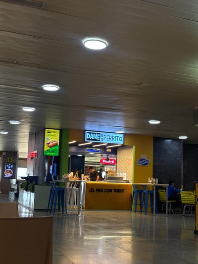 The image shows an indoor space, possibly an airport or large building hallway, with food vendors. "Dameunperrito," a hot dog vendor, is prominent. Nearby are "Plan-B" and "Plumrose" branding. People are visible at the vendors, likely customers. Seating arrangements with yellow and blue chairs are present. The overall lighting is bright with circular ceiling lights. The city is Catia La Mar, Venezuela.