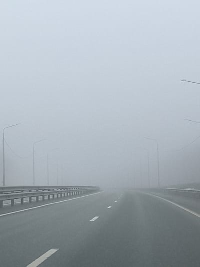 The image depicts a foggy highway in Сапуголи, Russia. The road surface is dark grey and marked with white lane lines. A metal guardrail runs along the left side of the road. Tall, indistinct streetlights are visible receding into the fog along both sides of the highway. The fog is dense, obscuring the distant view. There are no visible people or vehicles.