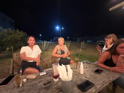 The image depicts a nighttime outdoor scene in Aljezur, Portugal, where four individuals are seated around a rustic wooden table.

From left to right:
A woman with dark hair, wearing a white collared shirt and dark shorts, is seated, looking towards the camera. Next to her, a woman with blonde hair, wearing a patterned sleeveless top and dark skirt, is seated, with her hands clasped, looking to her right. Further right, a young female with light brown hair, wearing a white top, is looking at a mobile phone she holds in her hands. On the far right, a young female with dark brown hair, wearing a dark sleeveless top, is partially visible, leaning her head on her hand and looking downwards.

The wooden table in the foreground holds various items: a dark mobile phone, a glass containing amber liquid, a light-colored wallet, a white rectangular card, a red bottle of ketchup, a small metal bucket filled with white paper napkins, a clear bottle of hand sanitizer, an empty glass, and two additional dark mobile phones.

The background is dark, illuminated by multiple streetlights. Sparse vegetation, including what appear to be agave plants, is present. Distant structures, likely buildings, and several parked vehicles are faintly visible under the artificial light. The ground is unpaved, consistent with a natural or semi-natural outdoor area.