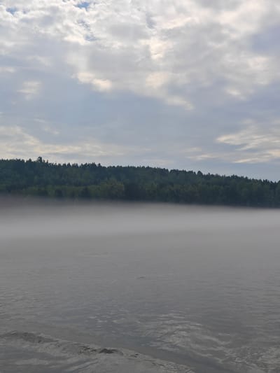 An overview of a misty body of water with a tree line in the background. The water is calm with ripples on the surface. A thick layer of fog hovers just above the water, obscuring the lower portion of the distant trees. The trees are lush and green, forming a dense forest along the horizon. The sky above is overcast with patchy clouds, some of which are illuminated by sunlight. The overall scene suggests an early morning or late evening in a natural setting. The location is the river in the village of Atamanovo, Krasnoyarsk Krai, Russia.