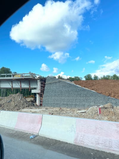 The image depicts a construction site in Novodrozhzhino, Russia. A newly constructed bridge is visible, with supporting steel and concrete structures. An excavator is parked nearby, surrounded by dirt and construction debris. Adjacent to the construction site is a retaining wall made of gabions, preventing soil erosion. The sky is blue with scattered clouds. A segment of a roadside curb with painted white and pink sections is present in the foreground.