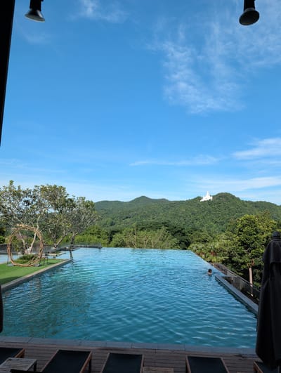 In Ban Pong Nuea, Thailand, an outdoor infinity pool overlooks a verdant, mountainous landscape. The pool's surface shimmers under a bright blue sky with scattered white clouds. A lone individual is partially submerged in the water on the right side of the pool. Lush green trees and foliage fill the midground, leading to rolling hills in the distance. A white building, possibly a temple or monument, is visible atop one of the prominent hills. The foreground features a wooden deck with lounge chairs, suggesting a resort or private residence. Two industrial-style lamps are visible in the upper left and right corners of the frame, attached to dark structures.